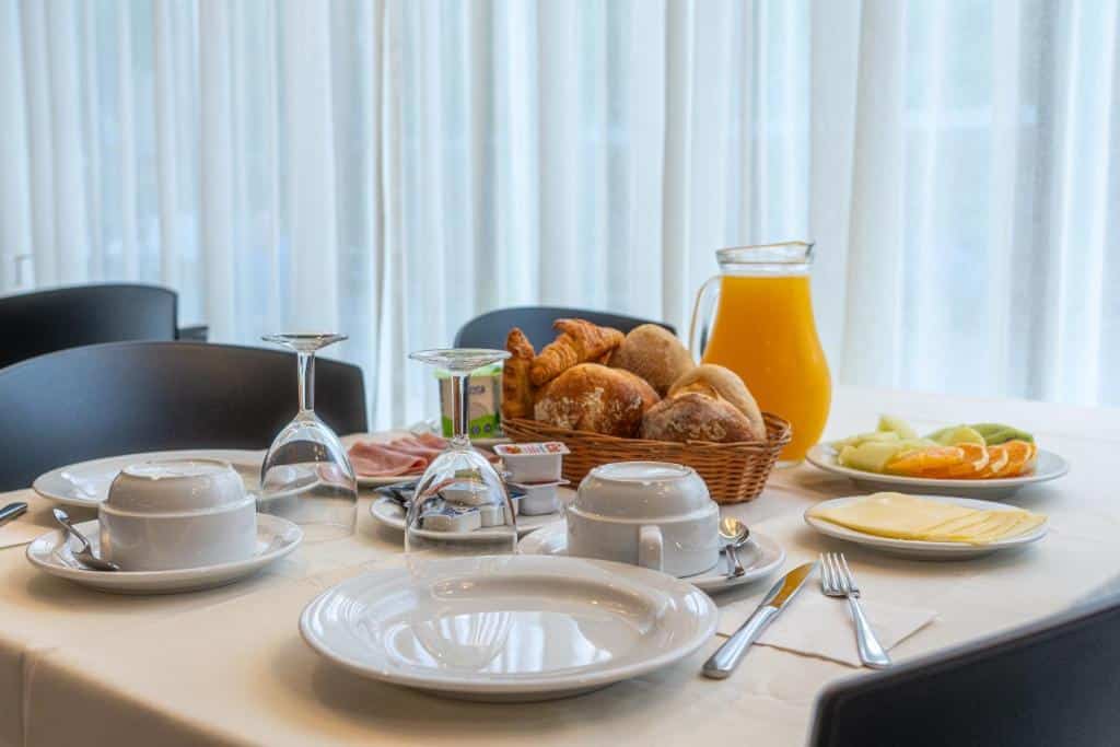 Mesa de pequeno-almoço com pão, fruta, queijo, sumo, pratos, chávenas, talheres e copos para duas pessoas, servida no Hotel Azinheira.