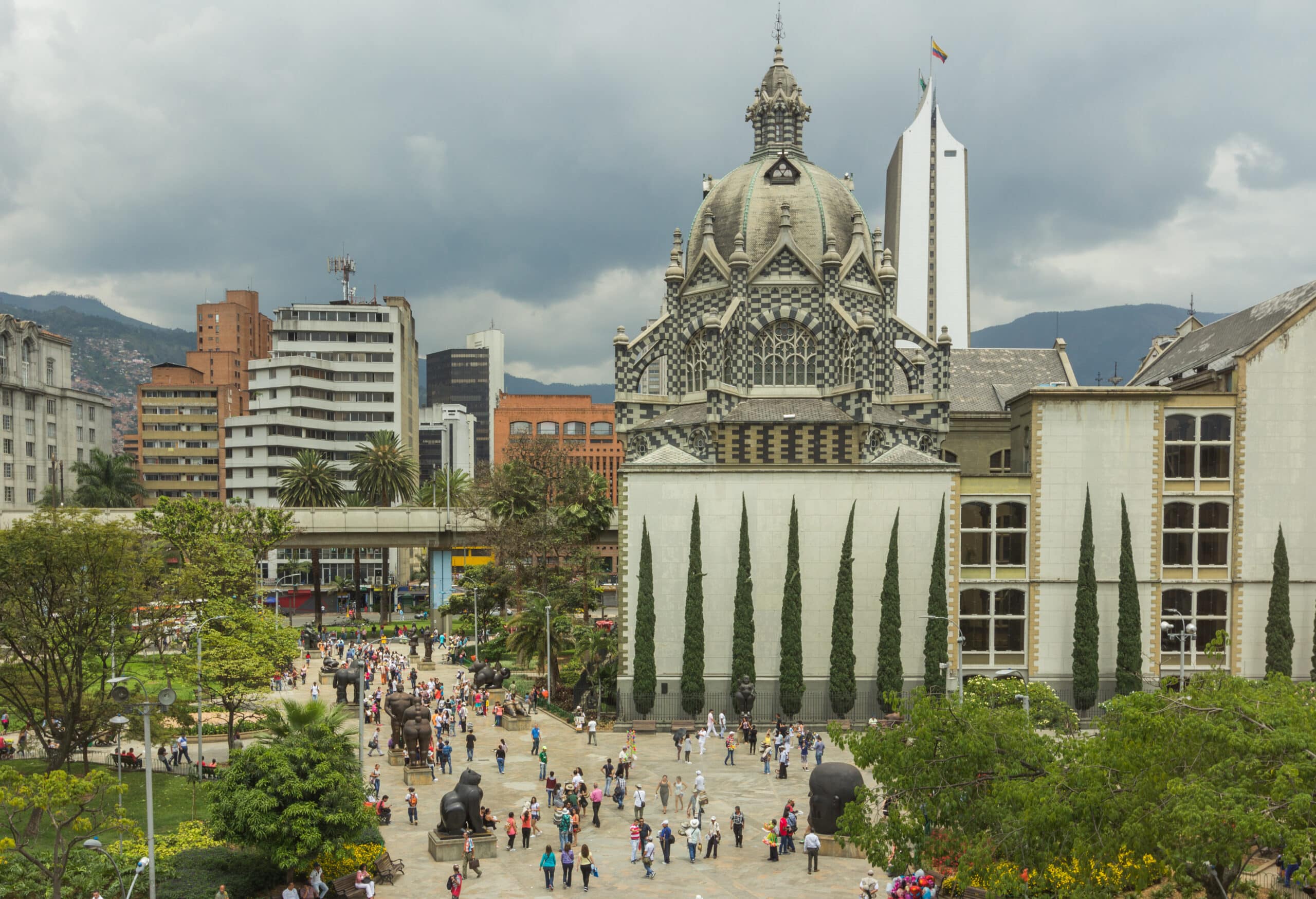 Pessoas a caminhar numa praça do centro de Medellín, com edifícios modernos e céu nublado ao fundo.