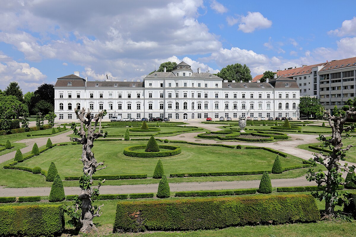 Palácio de Augarten, branco e com muitas janelas, visto por detrás de jardins geométricos bem cuidados, sob céu parcialmente nublado.