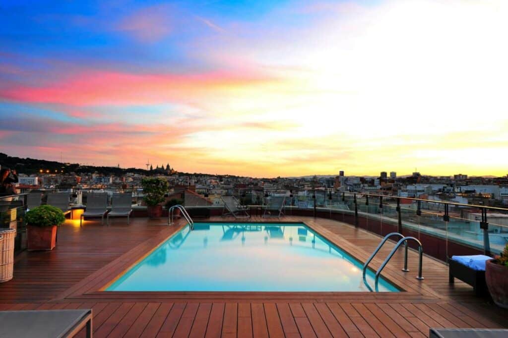 Piscina na cobertura do Hotel 1898, rodeada por um deque de madeira, plantas e espreguiçadeiras, com vista sobre a cidade ao entardecer.