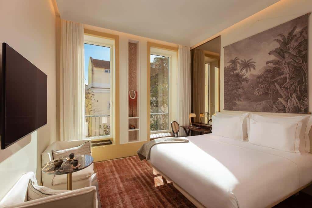 Quarto do The Ivens Autograph Collection, com duas janelas amplas com cortinas, um espelho com uma pequena mesa e cadeira, uma cama de casal com quatro almofadas e, na parede em frente, uma televisão com mesa e cadeiras estofadas