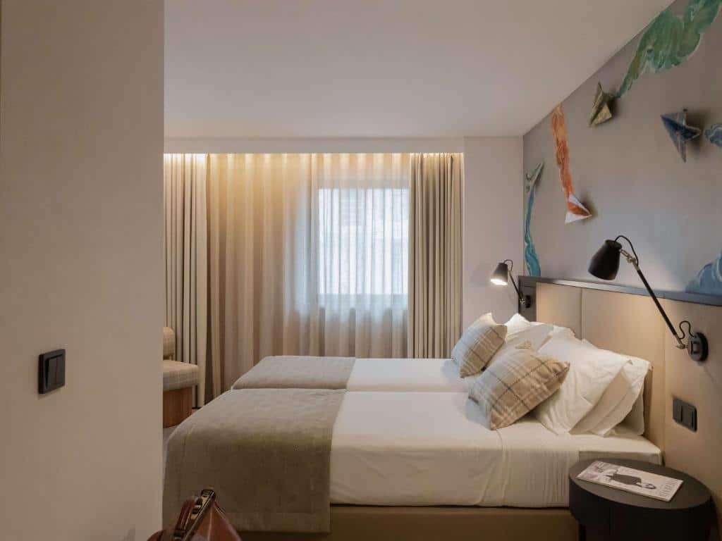 Quarto do Ibis Styles Lisboa Aeroporto com duas camas de solteiro, candeeiros de leitura na parede e janela com cortinas transparentes.