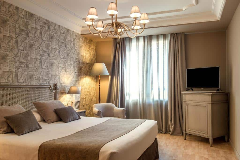 Quarto do Vincci Lys com ampla cama de casal, poltronas acolhedoras, candeeiros de mesa e lustre dourado no teto, com parede revestida a pedra natural em tons terrosos.
