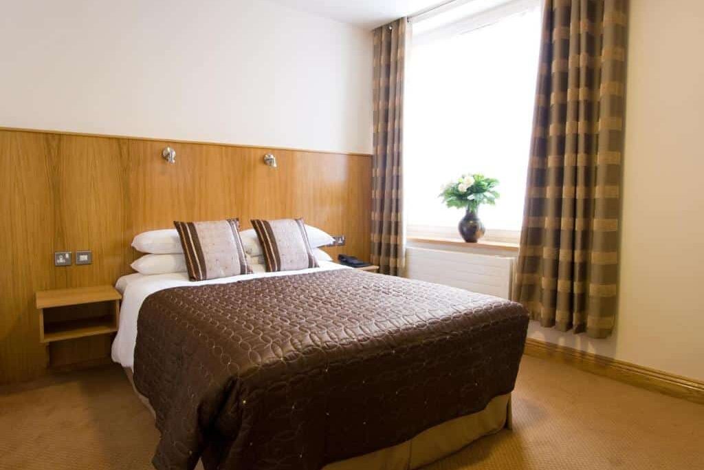 Quarto do Blooms Hotel com mobiliário de madeira, cortinas estampadas e um vaso de flores brancas à janela.