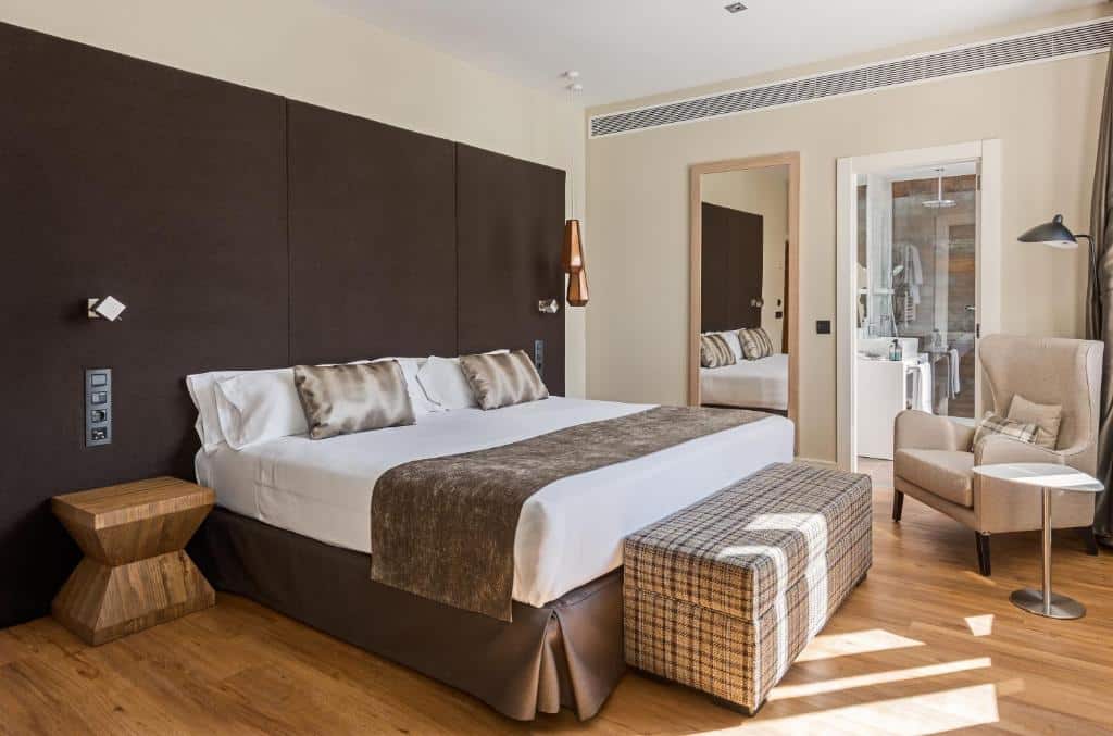 Quarto do Catalonia Gran Vía Madrid, um dos melhores hotéis em Madrid. A cama grande tem mesinhas de cabeceira dos dois lados e uma arca aos pés. Ao lado fica uma parede com espelho e a porta da casa de banho. Há uma poltrona com mesinha e candeeiro junto à porta.