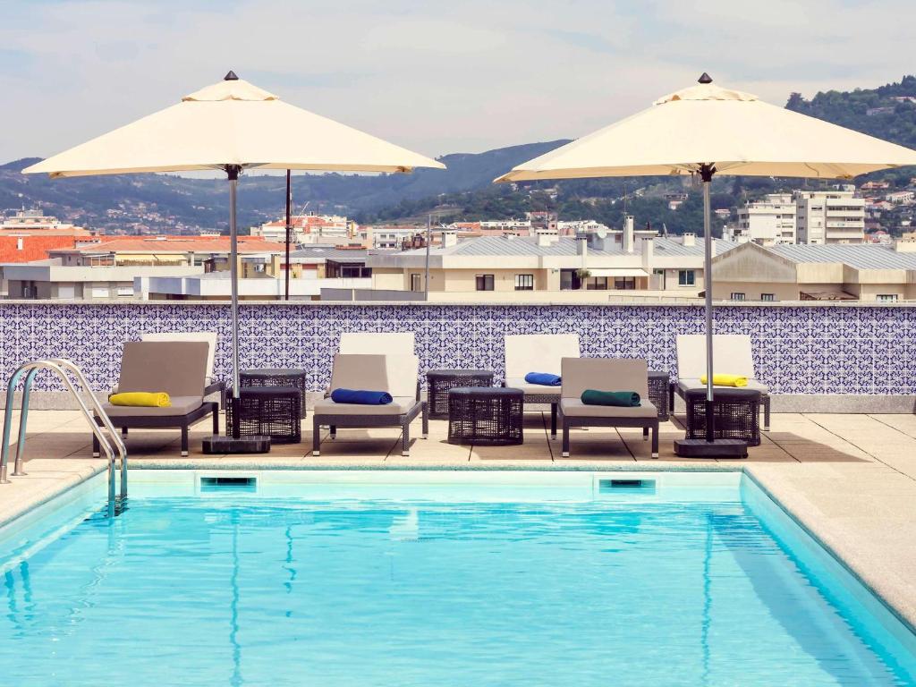 Piscina exterior do Hotel Mercure Braga Centro, com espreguiçadeiras, chapéus de sol e edifícios da cidade ao fundo num dia de sol.