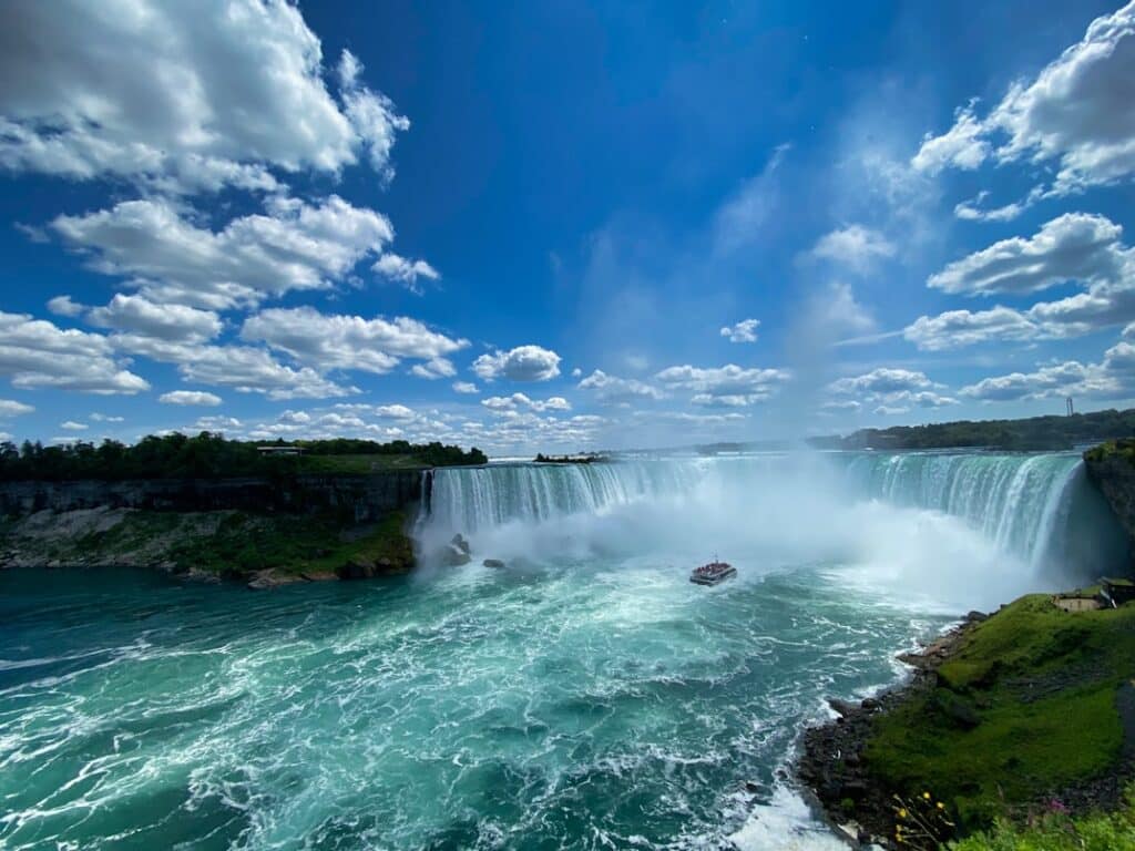 Cataratas do Niágara com névoa intensa e barco de turistas junto à borda, contra céu azul com nuvens brancas