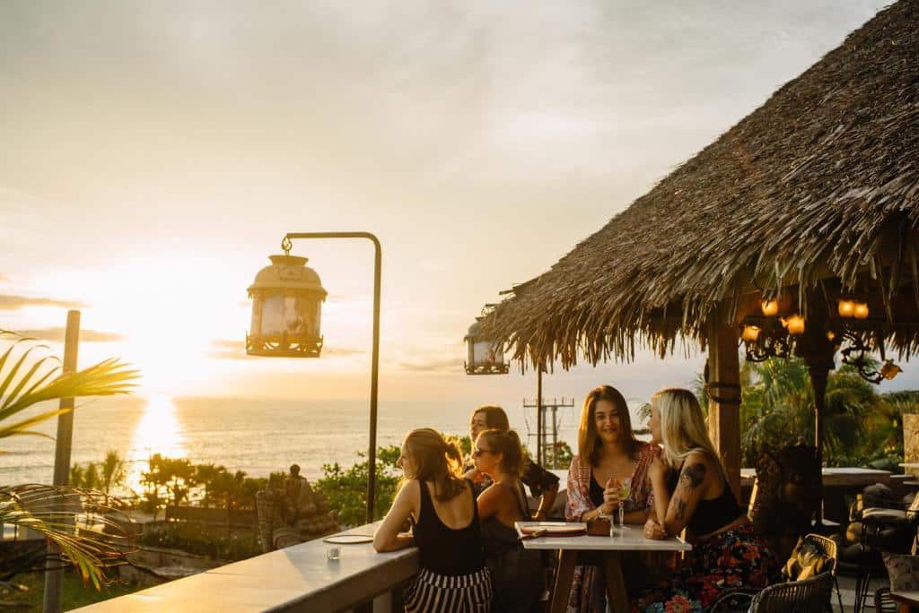 Grupo de pessoas num bar com teto de palha em Canggu, com vista para o pôr do sol sobre o oceano, em Bali.