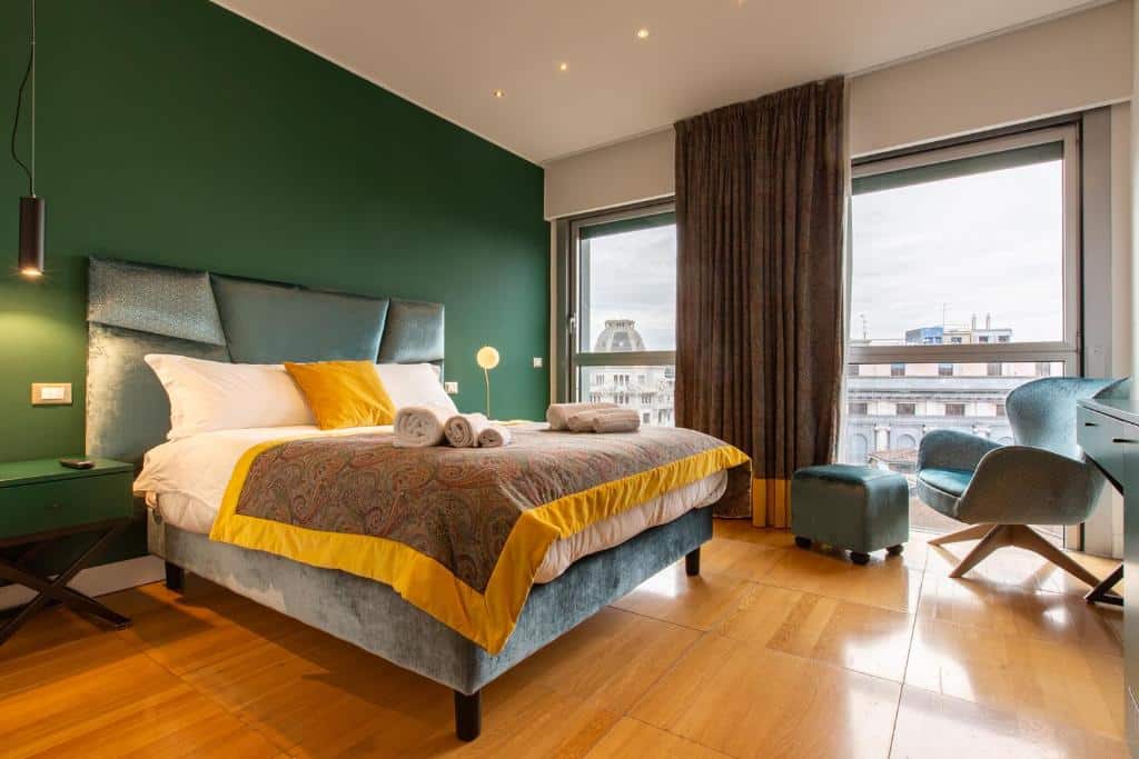 Quarto amplo do Missori Suites com duas janelas grandes com cortinas, uma cama de casal, uma poltrona e um pequeno bufete, além de duas mesas de cabeceira com candeeiros