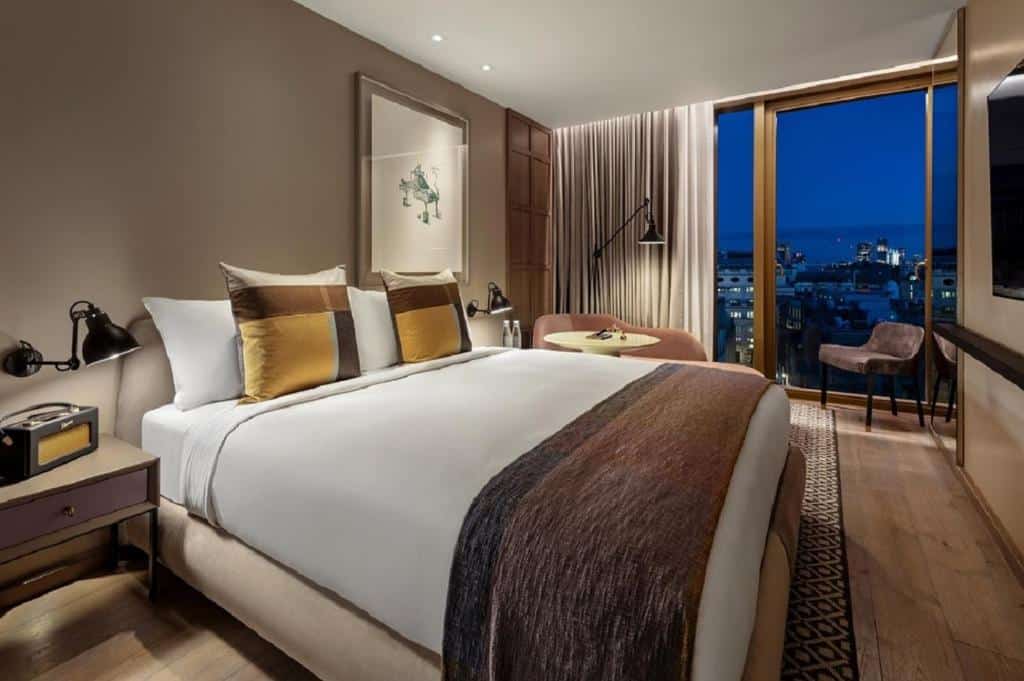 Quarto do The Londoner com cama de casal, uma janela panorâmica com cortinas, chão de madeira com tapete, duas mesas de cabeceira com candeeiros e uma poltrona.
