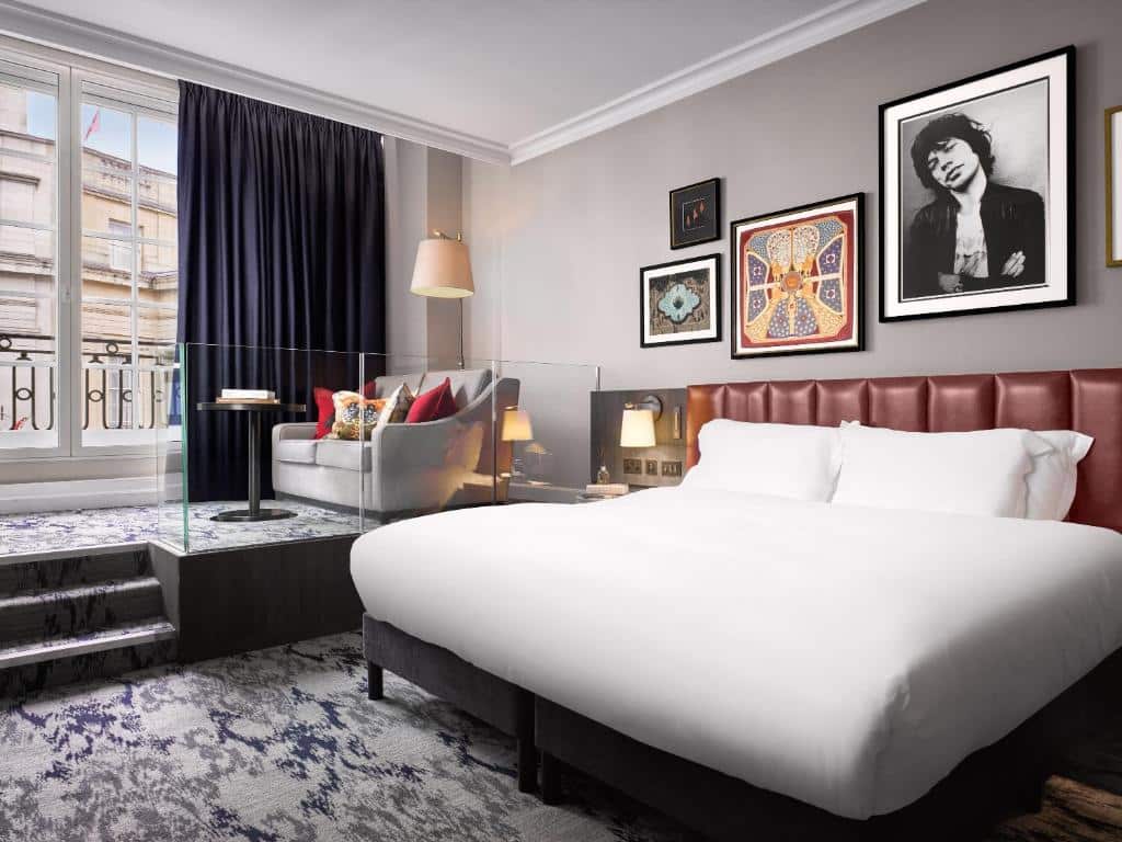 Quarto do The Trafalgar St. James com uma janela ampla com varanda e cortinas, um sofá com almofadas, um candeeiro, alcatifa, cama de casal com cabeceira em pele e alguns quadros na parede.