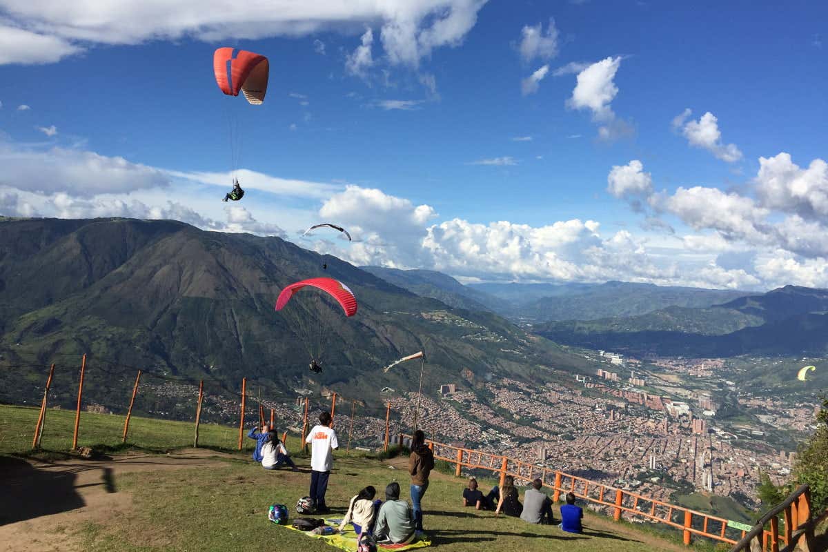 Pessoas sentadas numa colina relvada a observar parapentes a voar sobre a cidade e as montanhas, sob um céu azul com nuvens.