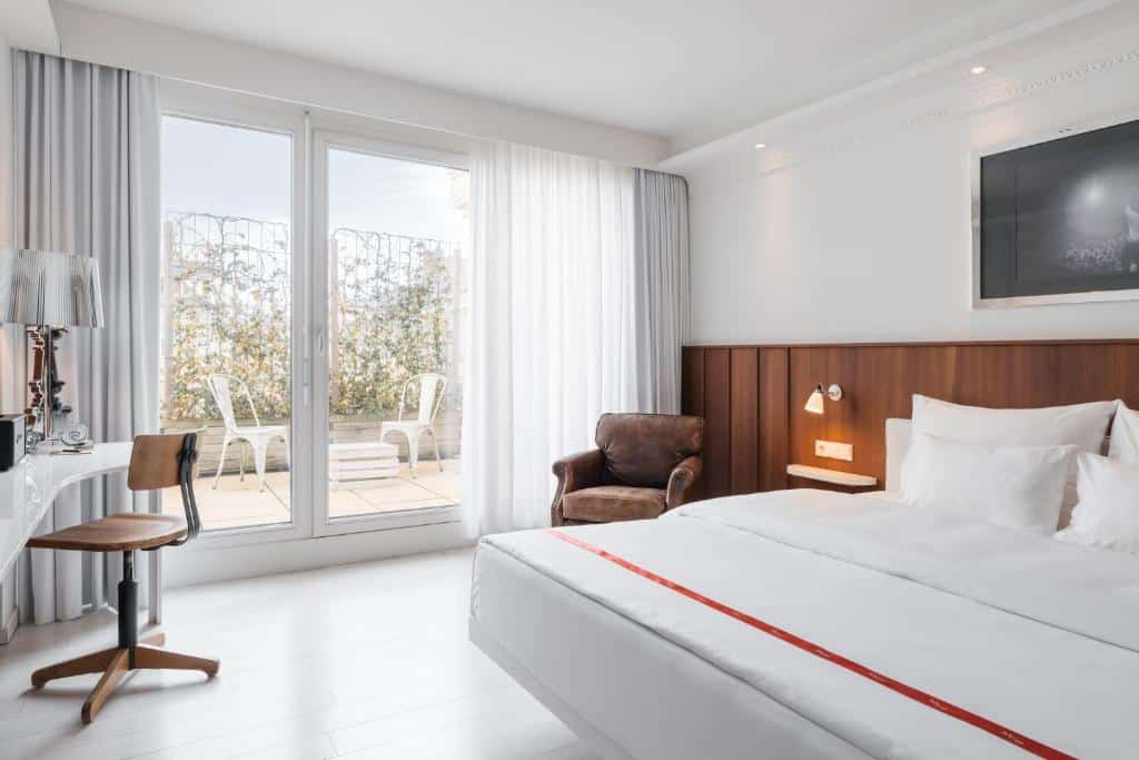 Quarto no Ruby Sofie Hotel, com cama de casal, secretária, cadeira, poltrona, TV e portas de vidro para um pátio com duas cadeiras.
