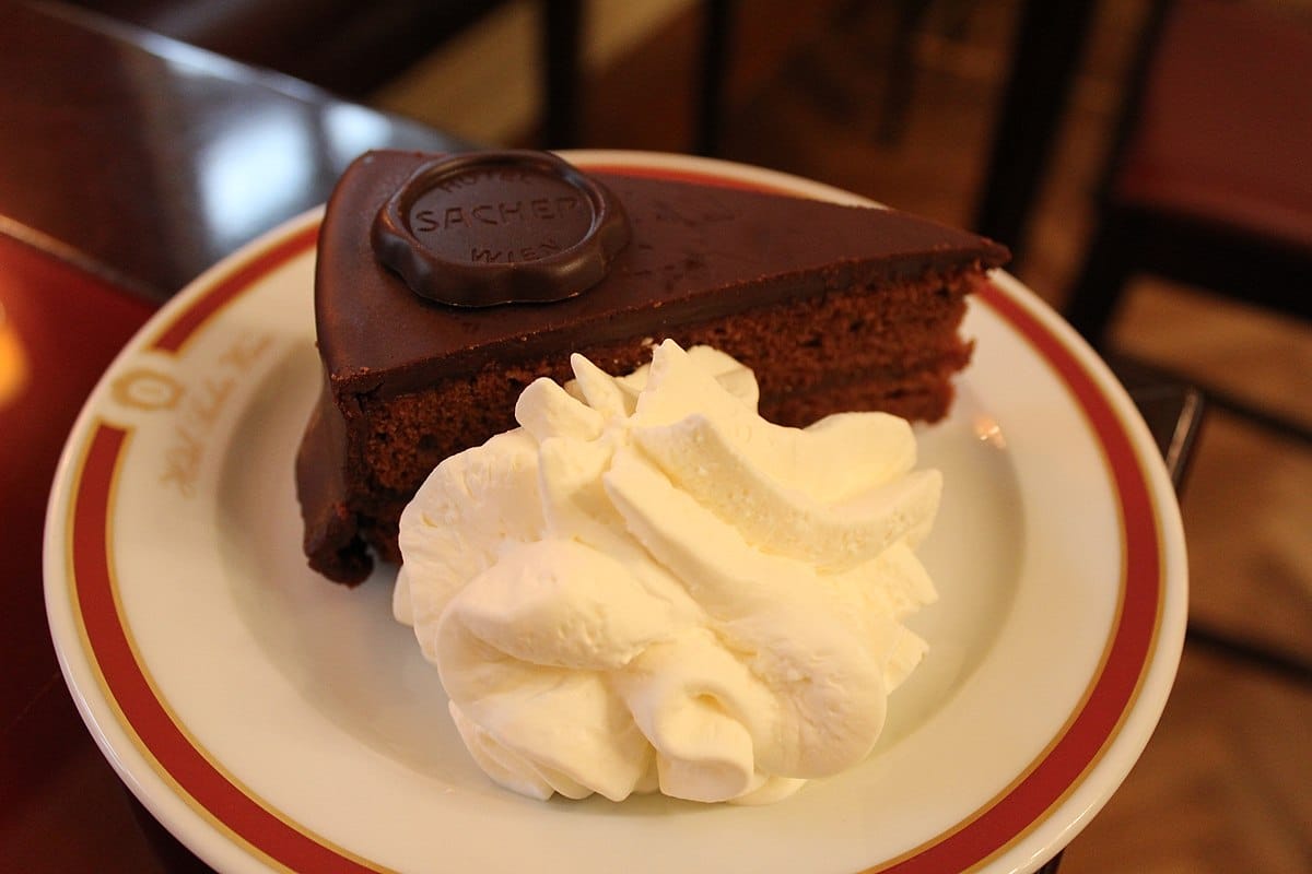Fatia de Sachertorte, bolo de chocolate com cobertura de chocolate, servida num prato com uma porção de natas batidas.