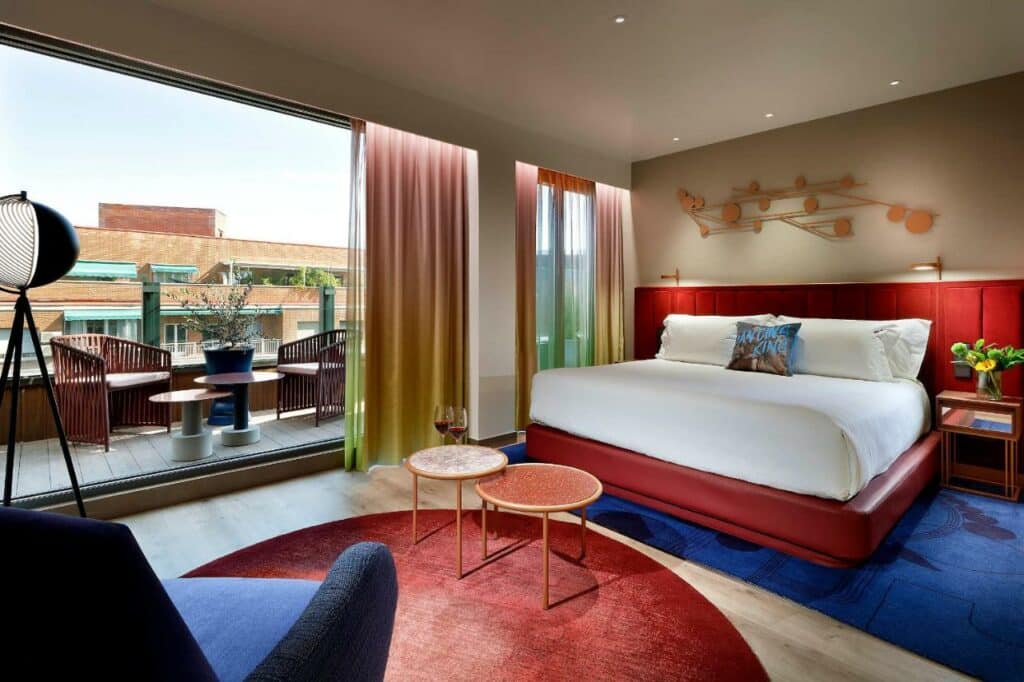 Quarto do Hard Rock Hotel Madrid, um dos melhores hotéis em Madrid. Uma ampla cama de casal está ao fundo do quarto e tem mesinhas de cabeceira e candeeiros verdes dos dois lados. Uma zona de estar em frente à cama tem mesinha, poltrona, candeeiro e tapete. A parede ao lado tem janelas amplas e uma porta de vidro que dá acesso à varanda com mesinha e cadeiras.