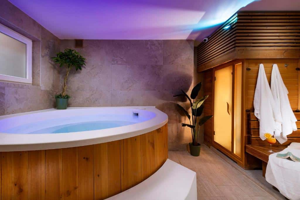 Banheira de hidromassagem redonda do Bo18 Hotel Superior ao lado de uma sauna de madeira, com dois vasos de plantas e roupões brancos pendurados numa sala de spa.