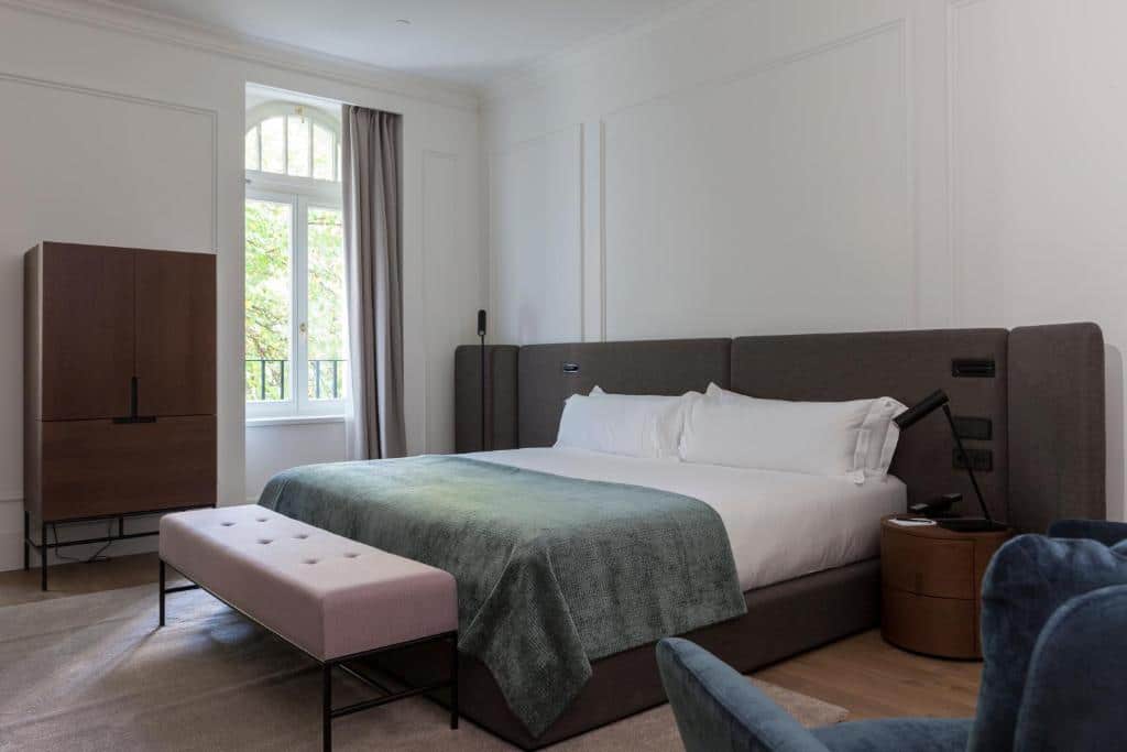 Quarto do Kozmo Hotel Suites &amp; Spa com uma cama grande, lençóis brancos, uma manta verde, um banco, um roupeiro e uma janela com cortinas.