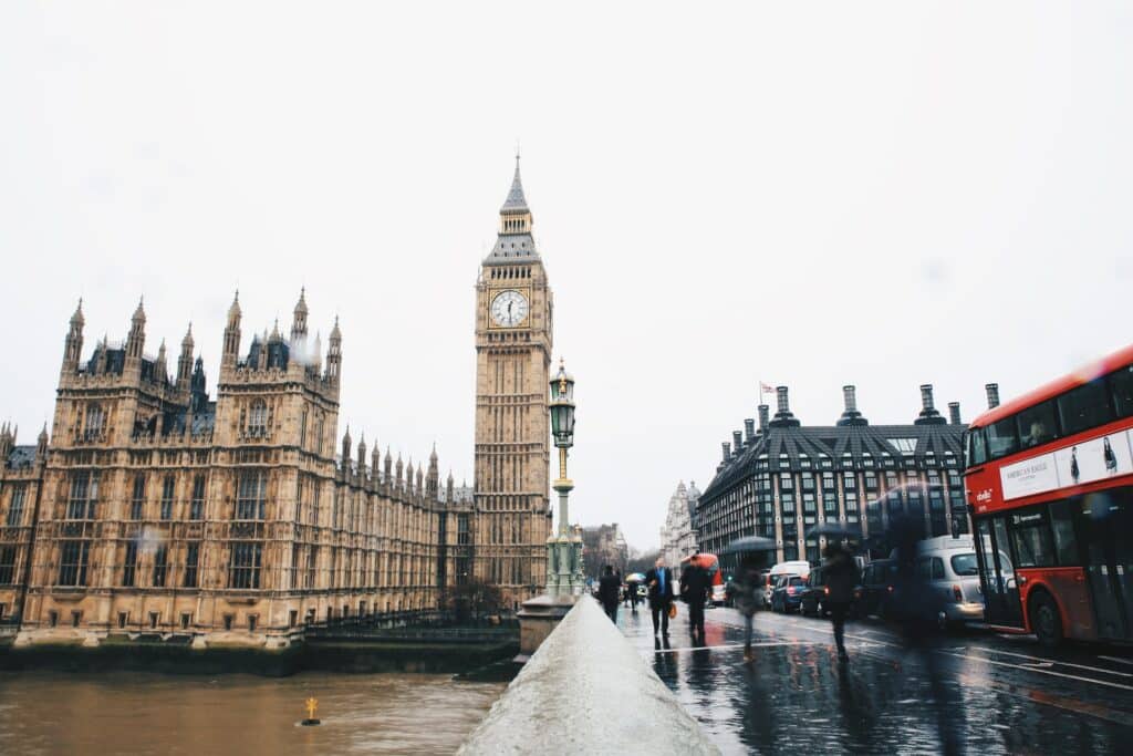 Edifício histórico com uma torre de relógio alta, o Big Ben, em frente ao rio Tamisa e a uma ponte movimentada, com autocarros, carros e pessoas a passar.
