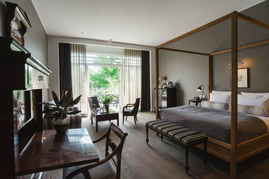 Quarto do Nimb Hotel com uma cama grande de casal com dossel e uma chaise longue. Tem também duas mesas, cadeiras, armário, vasos de plantas e uma varanda com cortinado.