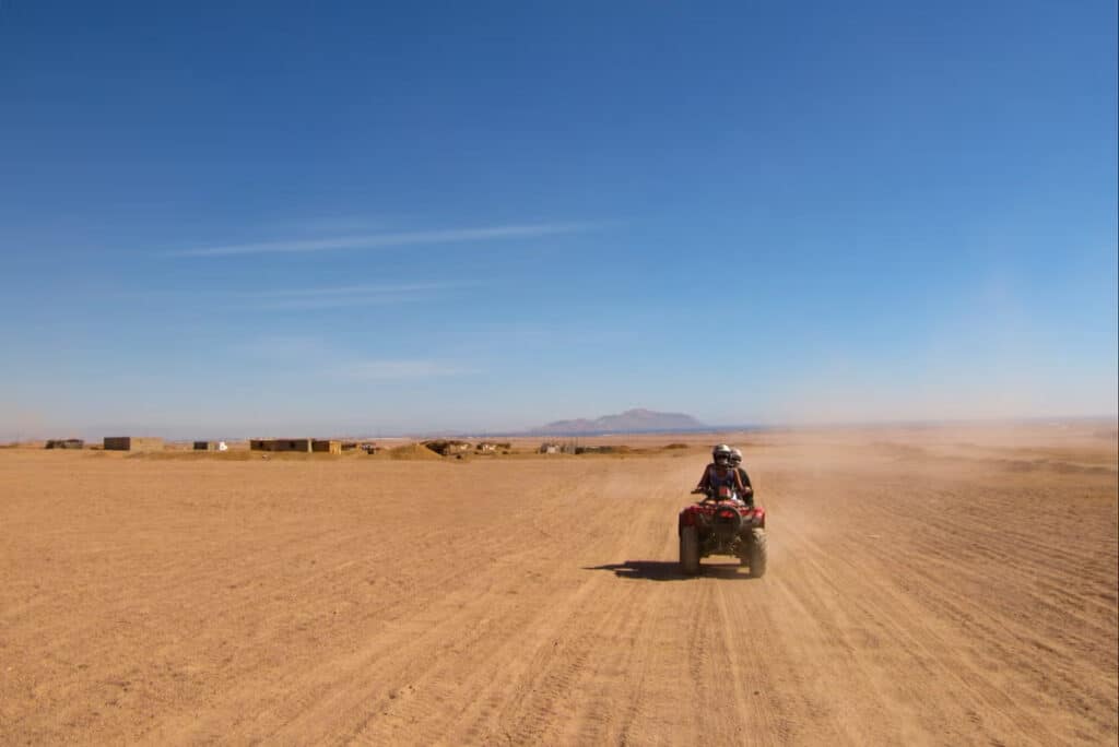 Uma pessoa numa moto-quatro atravessa uma vasta paisagem desértica junto a Agadir, sob um céu azul claro, com montanhas distantes no horizonte.