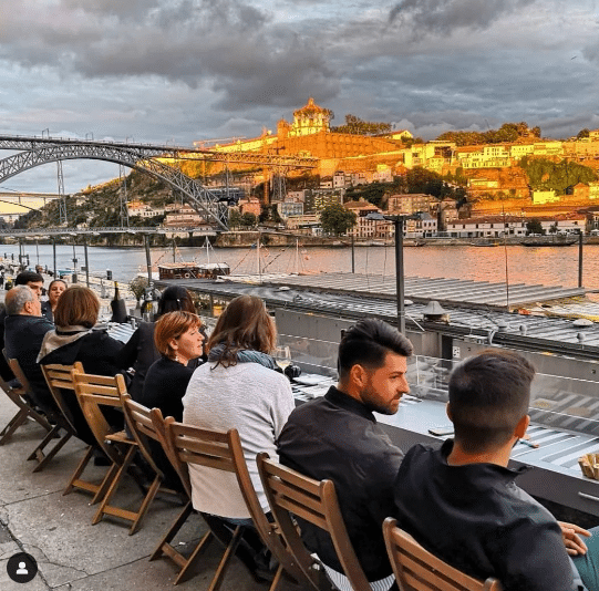 Pessoas sentadas no Wine Quay Bar ao ar livre, com vista para o rio, uma ponte e uma encosta histórica iluminada ao fundo.
