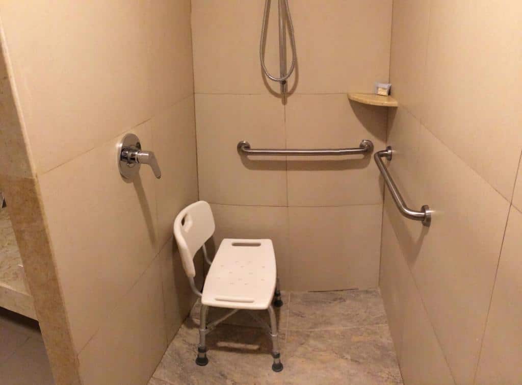 Chuveiro acessível com um assento branco, barras de apoio em duas paredes, um chuveiro de mão e uma pequena prateleira de canto.