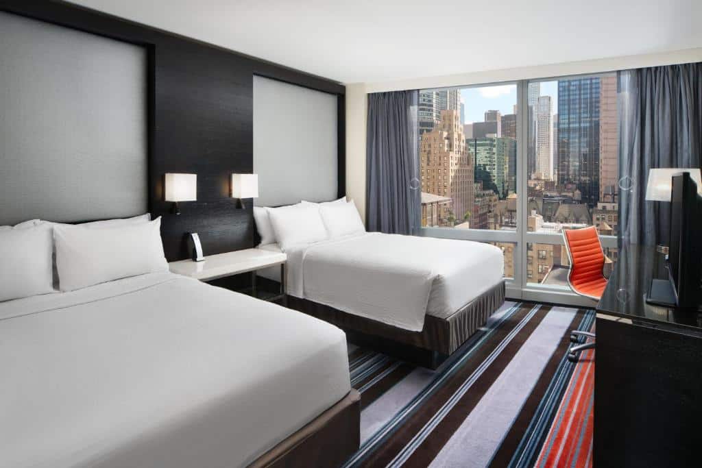 No quarto do Courtyard by Marriott New York Manhattan há duas camas de casal com roupa branca, lado a lado. À direita, uma secretária preta com cadeira de escritório laranja. Ao fundo, uma janela ampla com vista para os edifícios de Nova York.