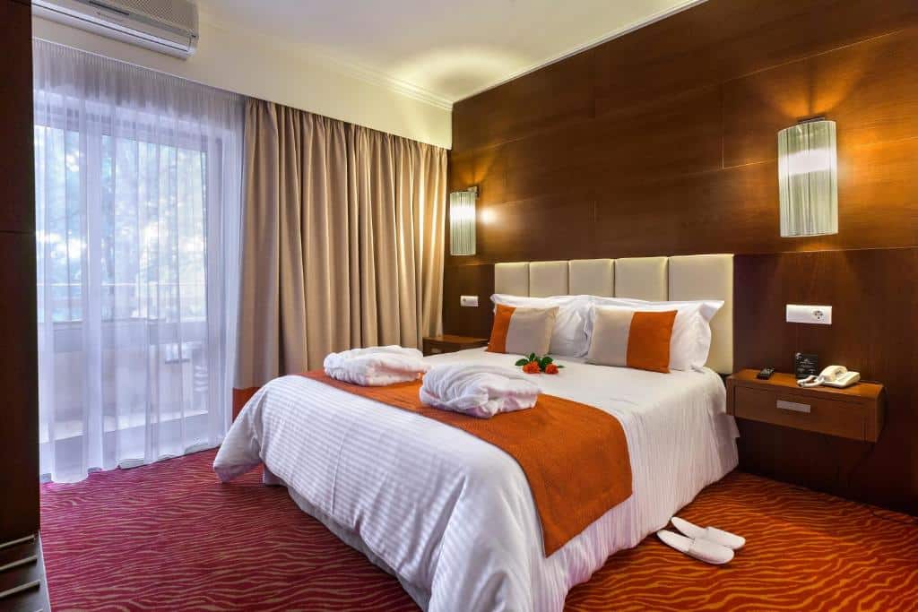 Um quarto na Avenida de Fátima com uma cama de casal, detalhes em laranja, dois roupões, mesas de cabeceira, candeeiros e uma janela com cortinas, para representar onde ficar em Fátima.