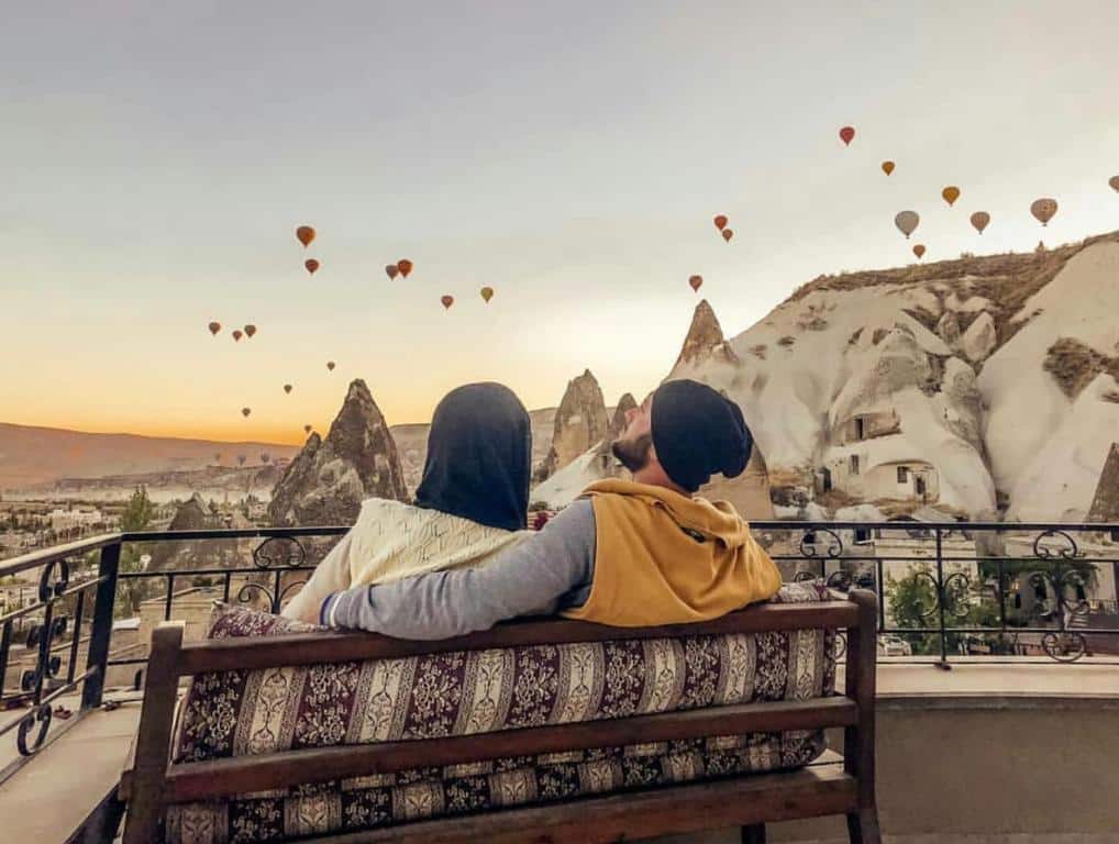 Um casal sentado num banco de frente para formações rochosas e balões de ar quente no céu ao amanhecer, no Elite Cave Suites.