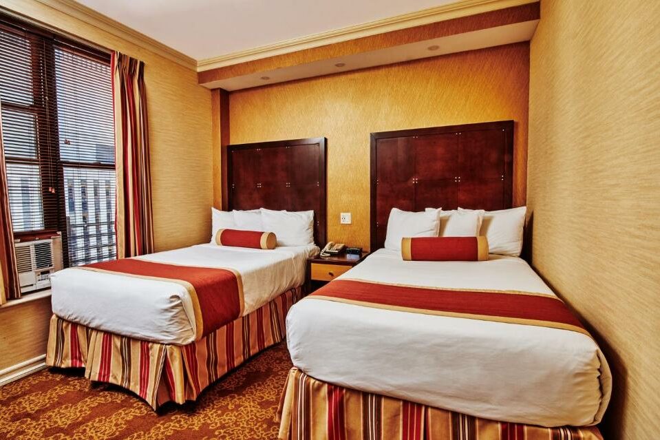 No quarto do Radio City Apartments há duas camas de solteiro com cabeceiras de madeira escura, lado a lado, cada uma com uma almofada cilíndrica vermelha. À esquerda, uma janela com estores. Decoração em tons bege com pormenores vermelhos e dourados.