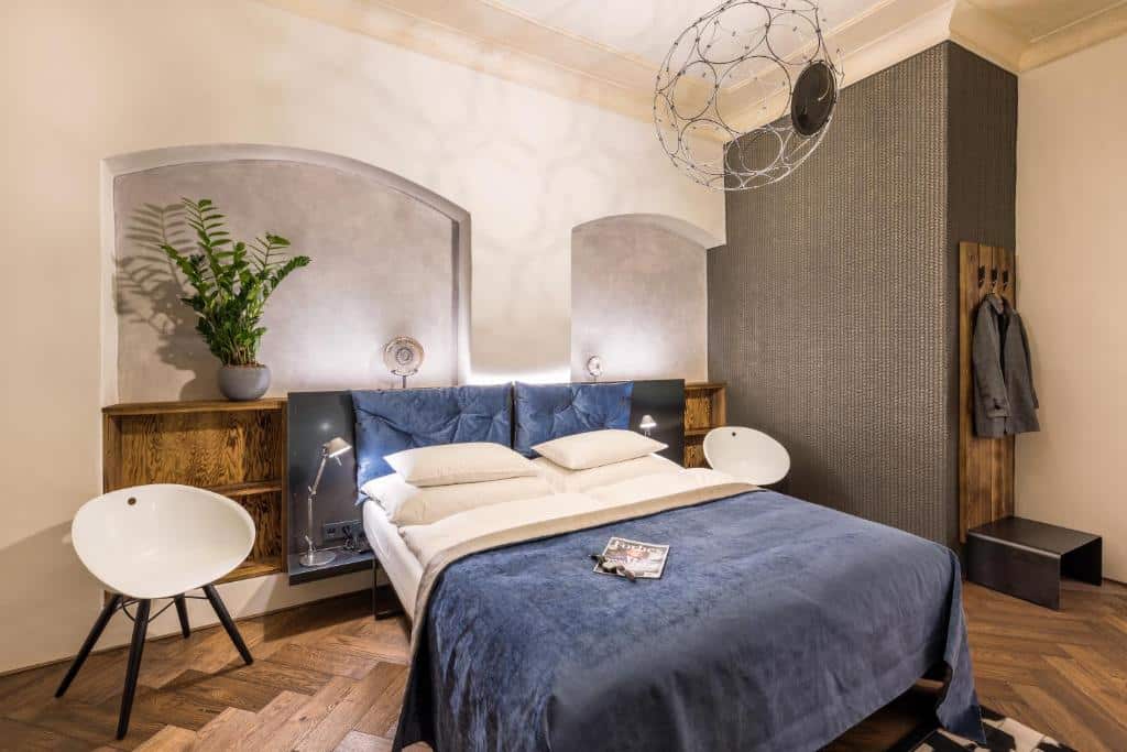 Quarto moderno do MOOo by the Castle, com cama de casal, colcha azul, prateleiras de madeira, duas cadeiras brancas e uma planta.