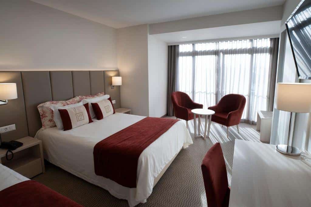 Um quarto no Hotel Fátima com uma cama de casal, duas cadeiras vermelhas, uma mesa redonda, candeeiros e uma secretária junto a grandes janelas com cortinas transparentes, para representar onde ficar em Fátima.