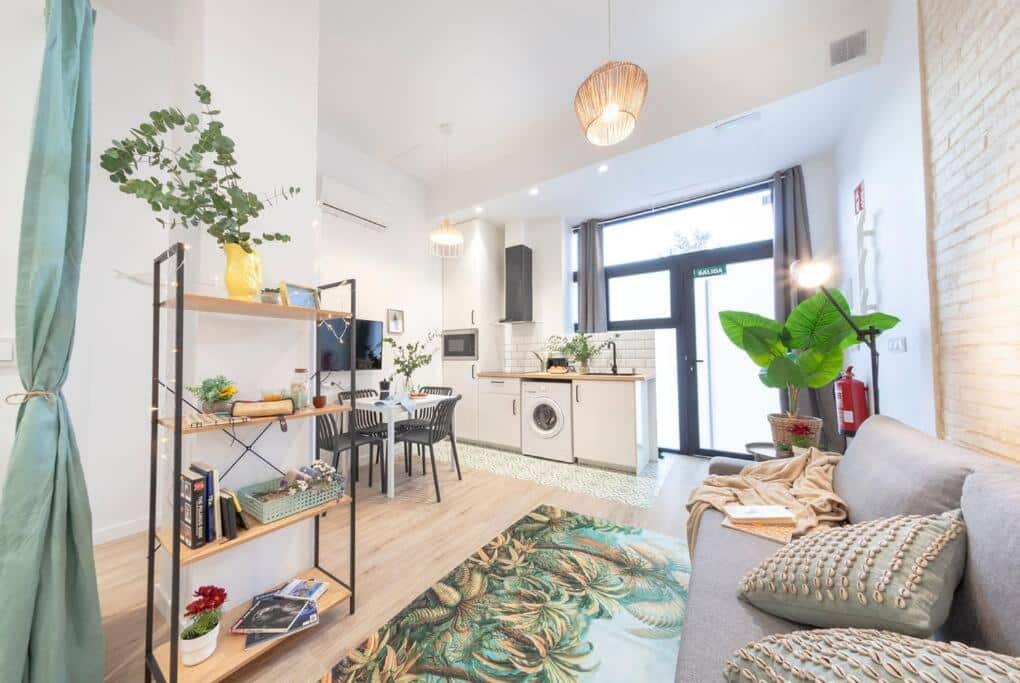 Sala de estar e cozinha integradas num apartamento moderno do AL5 Seaside Bliss – stylish loft, com decoração minimalista, luz natural, estante de madeira com plantas, sofá cinzento e tapete de estampa tropical.