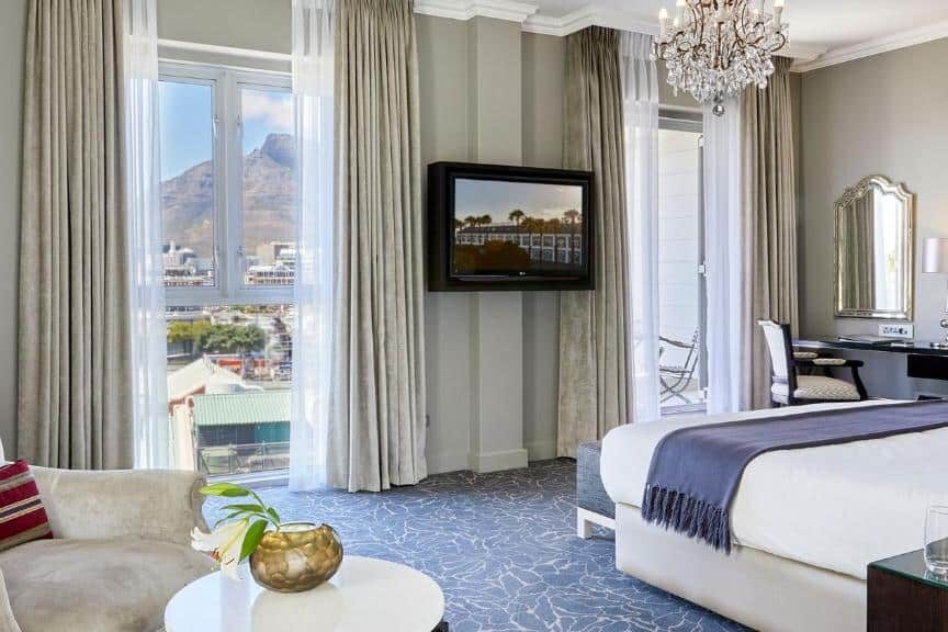 Quarto do Queen Victoria Hotel com cama, poltrona, secretária, TV e janelas com vista para a montanha e a cidade à luz do dia.