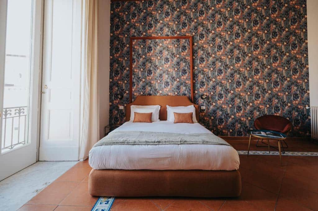 Quarto do Palazzo Pandola com cama de casal com dossel, parede de estampado floral escuro, janela com cortinas claras e poltrona a um canto.