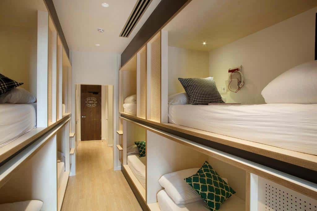 Quarto do TOC Hostel Barcelona. Tem beliches de ambos os lados, com almofadas, cobertor e tomada na parede, e uma porta ao fundo.