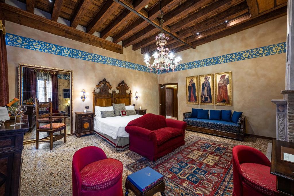Quarto luxuoso em estilo rústico com teto de madeira, paredes bege decoradas com ornamentos azuis e vermelhos, cama com cabeceira entalhada, sofá vermelho, tapete multicolorido e candelabro ornamentado