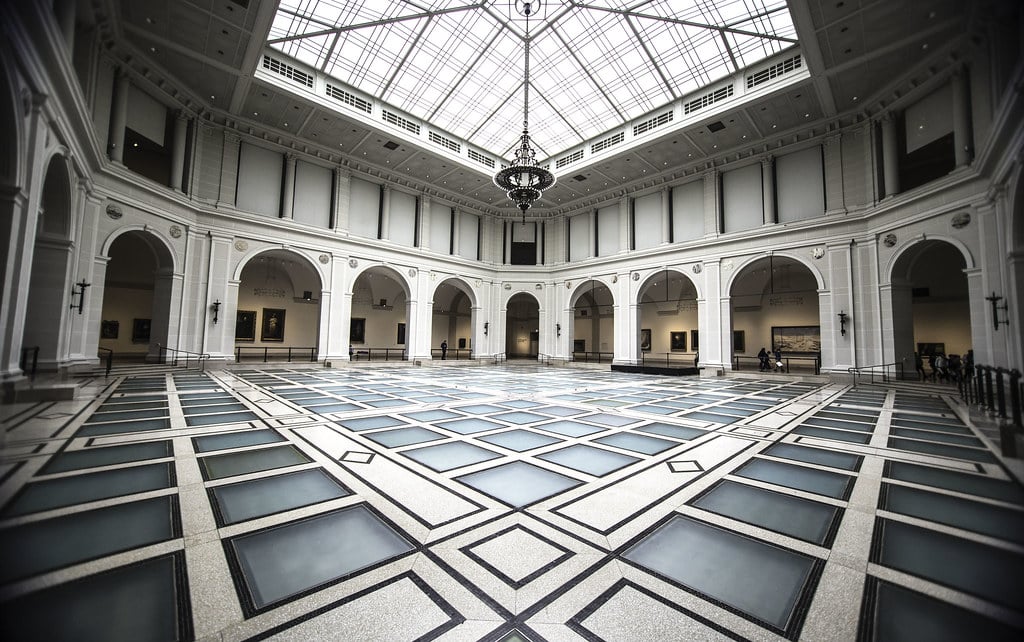 Átrio do Brooklyn Museum, com teto de vidro, pavimento de padrões geométricos em tons de azul e cinza e portas em arco.