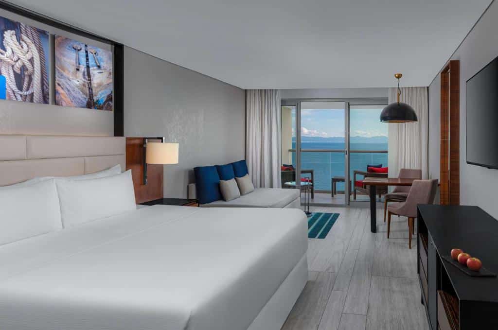 Quarto do Hilton Vallarta Riviera com uma cama king-size, zona de estar, secretária, janela grande e varanda com vista para o oceano.