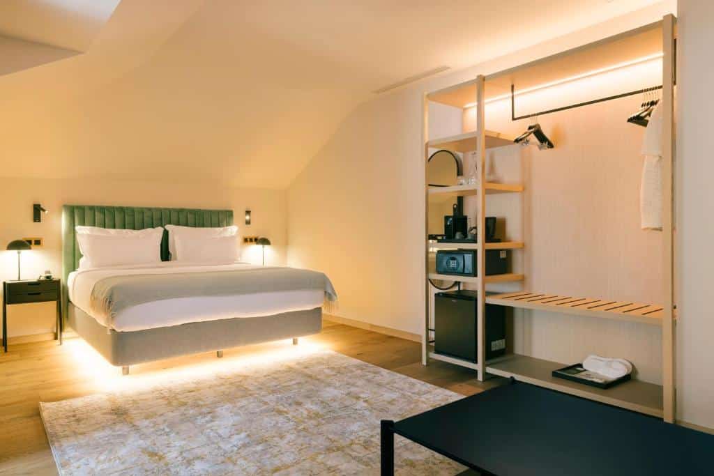 Quarto espaçoso do Pestana Rua Augusta Lisboa com uma cama de casal, mesas de cabeceira de cada lado com candeeiro, um armário aberto com cabides, sofá, conjunto para preparar café e um frigorífico, além de um tapete