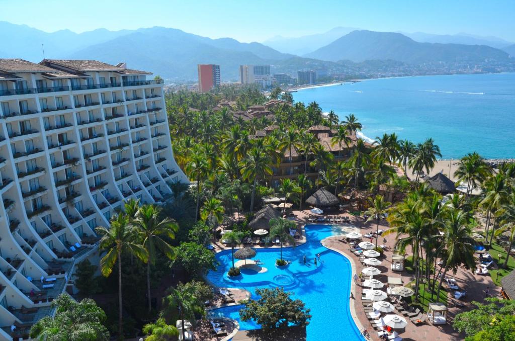 Um grande resort à beira-mar com um edifício curvo, piscinas, palmeiras e vista para o mar com montanhas distantes.