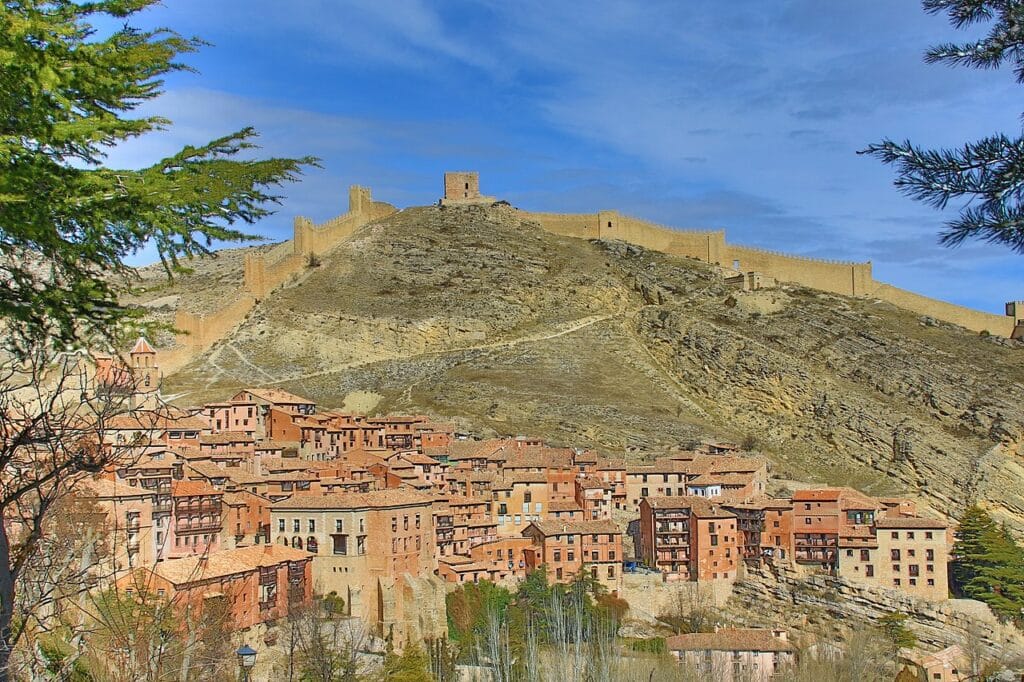 Vista de Albarracín, antiga vila espanhola e um dos destinos invulgares da Europa, encrustada nas colinas, com edifícios de tijolo vermelho e telhados de cerâmica. Acima da vila, estendendo-se ao longo do topo de uma montanha árida, há uma grande muralha de pedra com várias torres de vigia. O céu está limpo e há árvores verdes em primeiro plano, a emoldurar a cena.