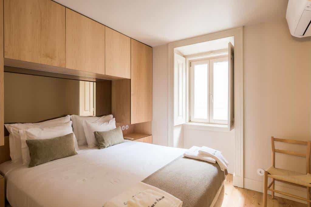 Quarto bem iluminado das Casas na Portuguesa com cama de casal, lençóis brancos, armários de madeira clara, uma janela, uma cadeira de madeira e um aparelho de ar condicionado.