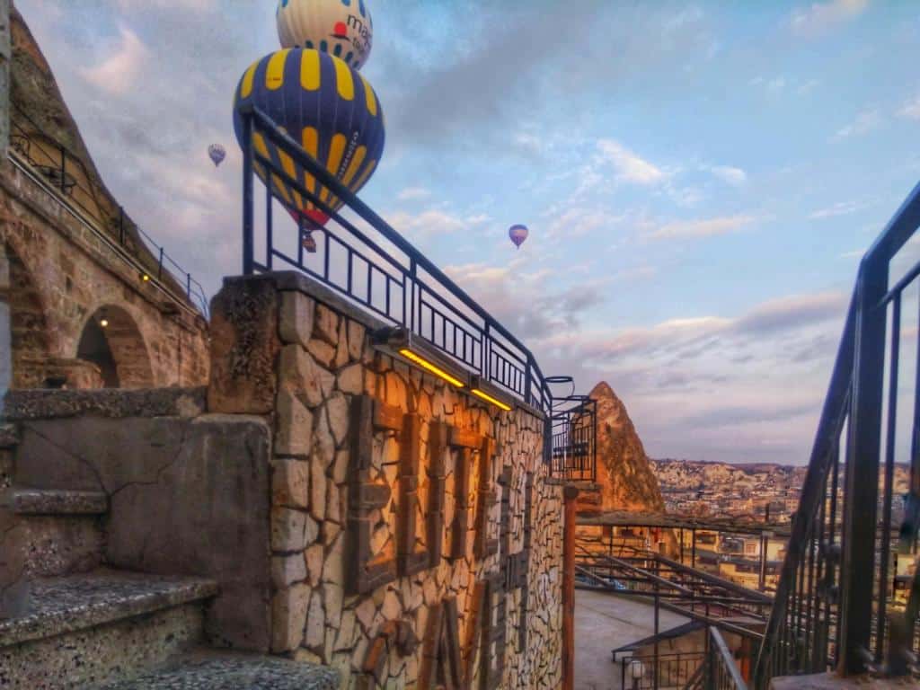 Edifício de pedra do Elite Cave Suites, um dos hotéis da Capadócia, com escadas e corrimão em primeiro plano; balões de ar quente e formações rochosas ao fundo sob um céu azul.