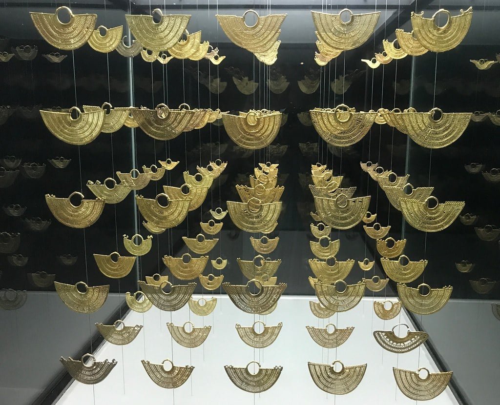 Peças de ouro em forma de leque, intrincadamente trabalhadas, suspensas por fios finos contra um fundo escuro, no Museu do Ouro Zenú.