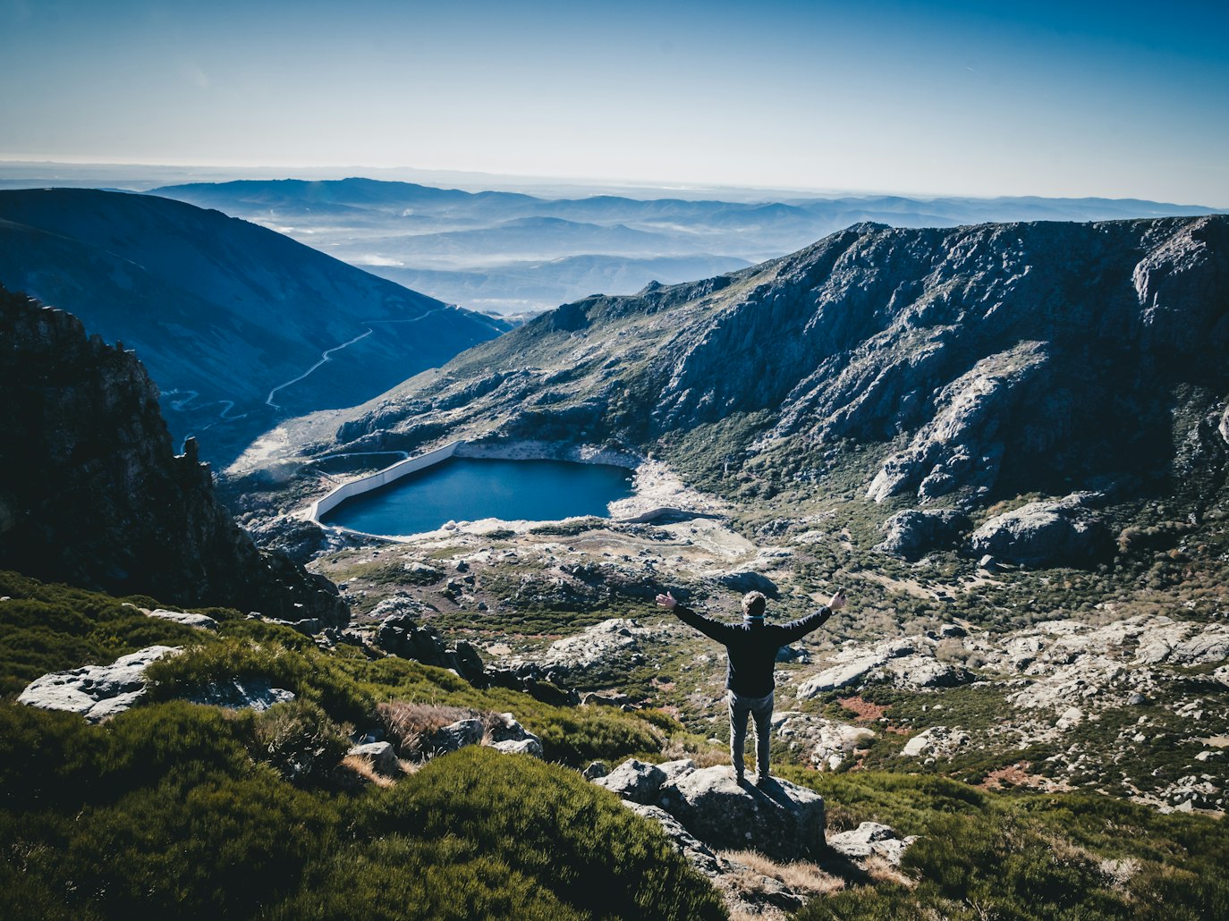 Pessoa de pé sobre uma rocha, de braços abertos, a observar uma paisagem montanhosa com um lago e colinas distantes na Serra da Estrela.