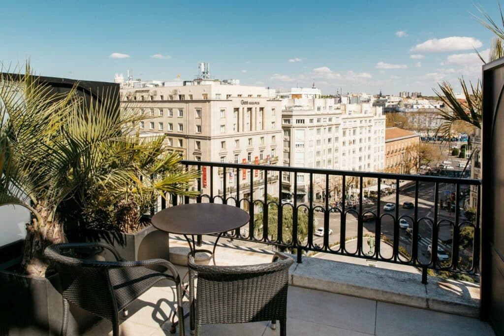 Terraço de um quarto no Hotel Villa Real. Com plantas dos dois lados e uma mesinha com cadeiras, tem grades em frente e vista do alto para a cidade.
