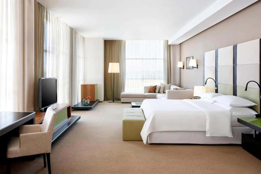 Quarto espaçoso do Sheraton Milan Malpensa Airport com cama de casal, sofá em L com almofadas, janelas amplas com cortinas e móvel com televisão