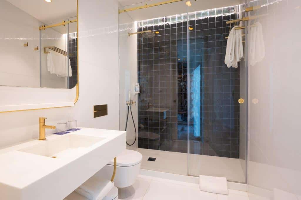 Casa de banho do Blue Liberdade Hotel com lavatório branco, base de duche em vidro, sanita branca e pormenores dourados, espelho amplo e toalhas brancas