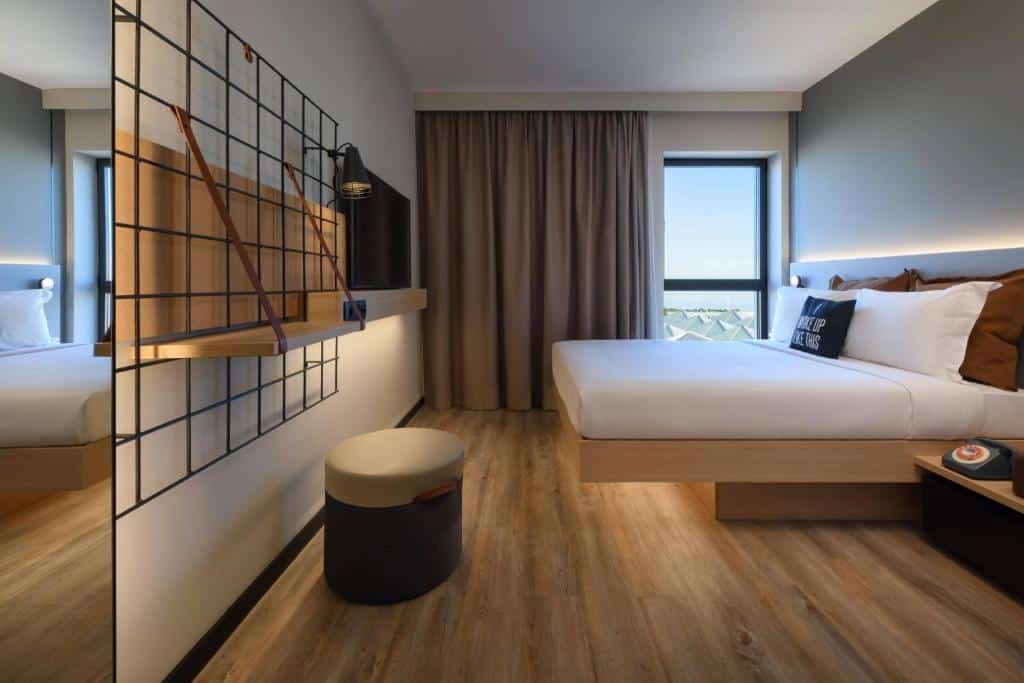 Quarto do Moxy Lisboa Oriente com uma janela com cortinas, uma cama de casal com almofadas, uma televisão em frente e, ao lado, uma pequena estante e um aparador.